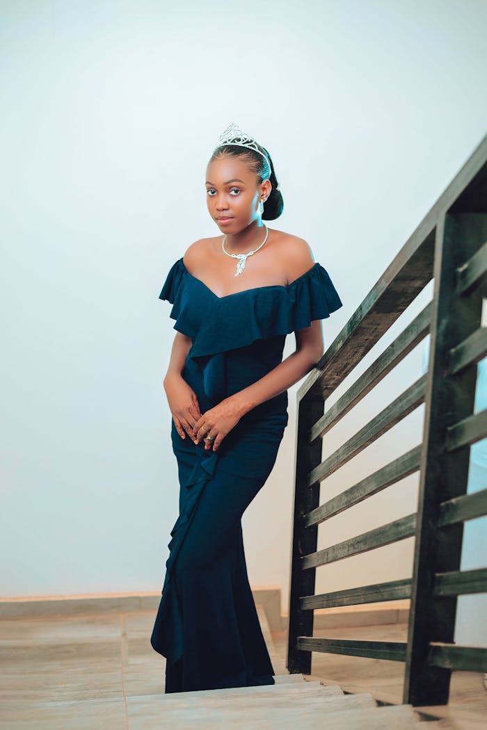 A young woman wearing a dark evening gown and tiara poses elegantly by a staircase.